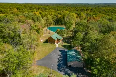 Aerial view of a log cabin with green roofing surrounded by lush trees with a private pond and a curving asphalt driveway in a secluded forest area.