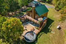 Aerial view of a two-story house with a large deck, surrounded by trees, featuring a trampoline and a propane tank in the yard.