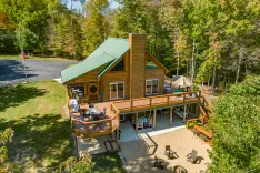 Aerial view of a two-story cabin with a green metal roof, large deck, and outdoor furniture surrounded by trees with a driveway and American flag on the left.