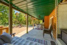 A cozy covered porch with green metal roof overlooking a sunny backyard with Adirondack chairs around a fire pit, adjacent to a wooded area.