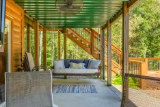 Cozy covered porch with a swing bed, cushioned chairs, and a multi-colored rug, overlooking a lush woodland area.