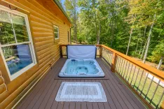 Spacious wooden deck with a hot tub and a white rug, surrounded by lush green trees in a serene forest setting.