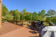 Spacious wooden deck of a cabin with outdoor furniture overlooking a forest with green trees under a clear blue sky.