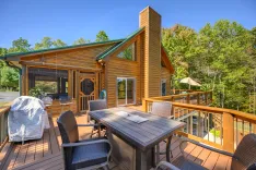 Spacious wooden deck of a log cabin with outdoor furniture and a barbecue grill, overlooking lush greenery.
