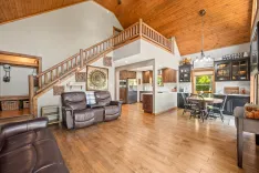 Spacious living room with high ceilings, wooden floor, leather sofas, and an open concept kitchen with modern appliances, leading to an upstairs loft area with wooden railing.