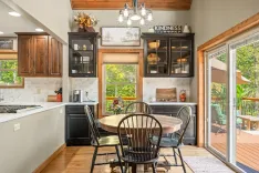 Modern kitchen interior with wooden cabinets, white subway tile backsplash, and a dining area with a round wooden table and chairs, leading to a deck with outdoor seating visible through glass doors. A sign reading "KINDNESS MATTERS" is displayed above the cabinets.