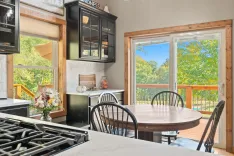 Bright and cozy kitchen interior with modern appliances, round wooden dining table, bouquet of flowers, and sliding glass doors leading to a sunny deck with a view of trees.