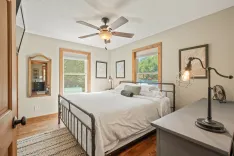 Bright and airy bedroom with a queen-sized bed, hardwood floors, ceiling fan, two windows with a view of greenery, and a decorative bedside lamp.