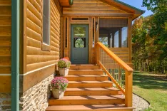 Wooden log cabin entrance with stairs, flower pots, and a screened porch surrounded by trees.
