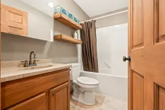 A modern bathroom with wooden cabinetry, granite countertop with undermount sink, two floating shelves above the toilet, and a shower-tub combo with a brown curtain.