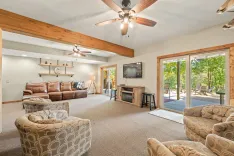 Spacious living room with natural light, featuring a large brown leather couch, patterned armchairs, wooden beams, a ceiling fan, fireplace, mounted television, and sliding glass doors leading to an outdoor area.