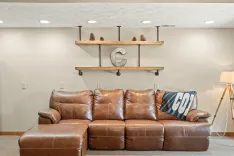 Alt: A cozy living room corner featuring a brown leather sectional sofa with a "GO" blanket thrown over it, industrial-style wooden shelves with decorative pine cones and a metal letter "C", and a wooden floor lamp to the side.