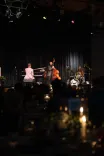 A dimly lit jazz club scene with musicians on stage performing in front of an audience, with focus on the pianist and bassist.