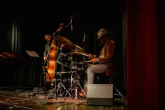 Two musicians on stage, one playing a double bass and the other playing a drum set, with a music stand and stage lighting in the background.
