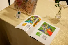 Open brochure with colorful artwork and QR codes on a table with a white rose in a vase and a hand sanitizer bottle.