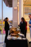 Two individuals conversing near a food table at an indoor event with windows showing a cityscape at dusk in the background.