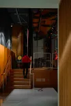 A person in a red coat walking up the stairs in a dimly lit corridor with industrial elements and warm wood paneling.