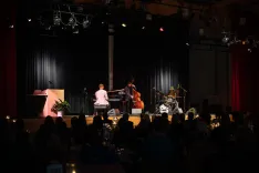 Alt text: A jazz trio performing on stage in a dimly lit venue with an audience in the foreground.