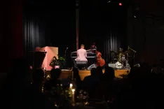 A dimly lit jazz club scene with silhouettes of a pianist and a bass player performing on stage, watched by an audience in the foreground.