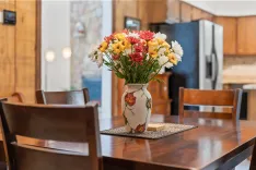 A colorful bouquet of flowers in a vase with painted leaves on a wooden dining table with chairs, in a kitchen setting with wooden cabinetry and a fridge in the background.