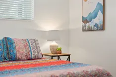 A cozy bedroom corner featuring a colorful patterned bedspread, a bedside table with a lamp and a small plant, and a framed mountainous landscape artwork on the wall.