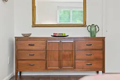 A wooden mid-century modern sideboard with drawers and cabinets decorated with a bowl, a plate with colorful mugs and a large green vase, positioned below a gold-framed mirror opposite a window with white blinds.