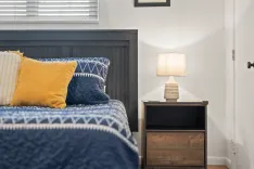 Cozy bedroom corner with a neatly made bed with blue bedding and a yellow accent pillow, beside a nightstand with a lamp.