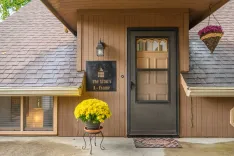 A cozy wooden house entrance with a brown door and welcome mat, a sign reading "The Artist's A-Frame", a hanging flower basket, and a pot of bright yellow chrysanthemums.