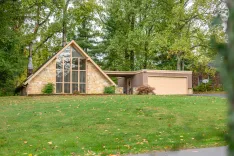 A mid-century modern house with a large front gable window, stone facade, and attached garage surrounded by trees in a suburban setting.