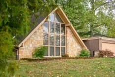 A modern A-frame house featuring large triangular windows and stone wall accents surrounded by a green lawn with scattered autumn leaves.
