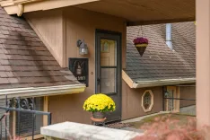 Exterior view of a brown house with a black front door, a hanging flower basket, and a large potted yellow chrysanthemum by the entrance.
