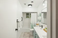 A modern, bright bathroom with a bathtub and shower curtain, a toilet, a vanity with a marble countertop and two sinks, a large mirror, and wall decorations with plant motifs.