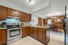 Modern kitchen interior with wooden cabinets, stainless steel appliances, a mosaic backsplash, and an attached dining area with a wooden table set.