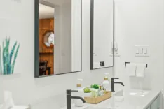 Modern bathroom interior with a white countertop, black faucet, hand soap dispensers, and a reflection of an adjoining room in the mirror.