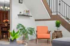 Cozy home interior with mid-century modern decor, featuring an orange accent chair, potted plants, decorative shelves, and a partial view of a dining area with a wooden table.