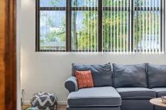 Modern living room corner with a gray sectional couch, decorative pillows, a small pouf, and a large window showing greenery outside.
