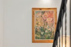 A framed impressionist-style painting of flowers hanging on a white wall, with part of a lampshade and blurred black objects in the foreground to the right.
