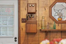 Vintage wooden wall-mounted telephone on a rustic wooden panel wall beside a window with blinds, next to an antique mirror and a shelf with old glass bottles.