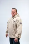 Man standing confidently wearing a beige hoodie with a circular logo on the chest and an American flag on the sleeve, against a light gray background.