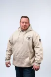 Man standing confidently in a beige hoodie with a circular logo on the chest and an American flag on the right sleeve, against a light background.