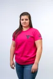 Young woman standing against a light background wearing a bright pink t-shirt with a graphic design on the chest and left sleeve, paired with blue jeans.