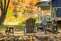 Three Adirondack chairs and a wooden stool on a rock-lined backyard patio with fallen leaves, near a house with a porch in an autumn setting.