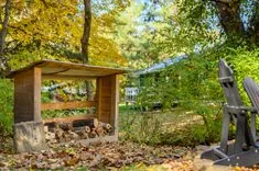 A wooden log shelter and Adirondack chair in a backyard with autumn leaves on the ground, with a blurry house in the background.