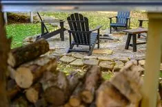 A cozy outdoor seating area with black Adirondack chairs and a table surrounded by autumn leaves and a stone border, viewed from behind a pile of firewood.
