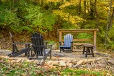 Two Adirondack chairs and a wooden table surrounded by autumn leaves in a forest setting.