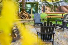 Outdoor scene with black Adirondack chairs and a wooden picnic table surrounded by autumn leaves, blurred yellow foliage in the foreground, and a stack of firewood in the background.