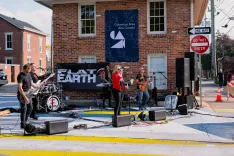 A band performing live on a street stage with a banner reading 'Flat Earth' in the background.