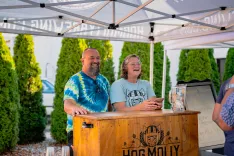 Two smiling people standing behind a wooden bar under a tent with the sign "HOG MOLLY" at an outdoor event.