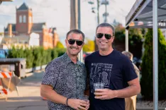 Two smiling men wearing sunglasses posing for a photo with a beverage, with urban scenery in the background.