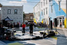 Outdoor street concert with musicians performing in front of a casual audience, surrounded by buildings with colorful murals.